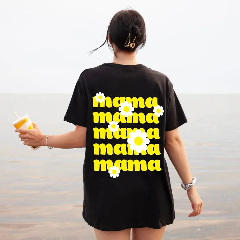 A woman stands by the water, holding a drink and wearing the Mama Daisy • Black Tee—a comfy unisex-fit Bella/Canvas shirt featuring “mama” in bold yellow with white daisies on the back.