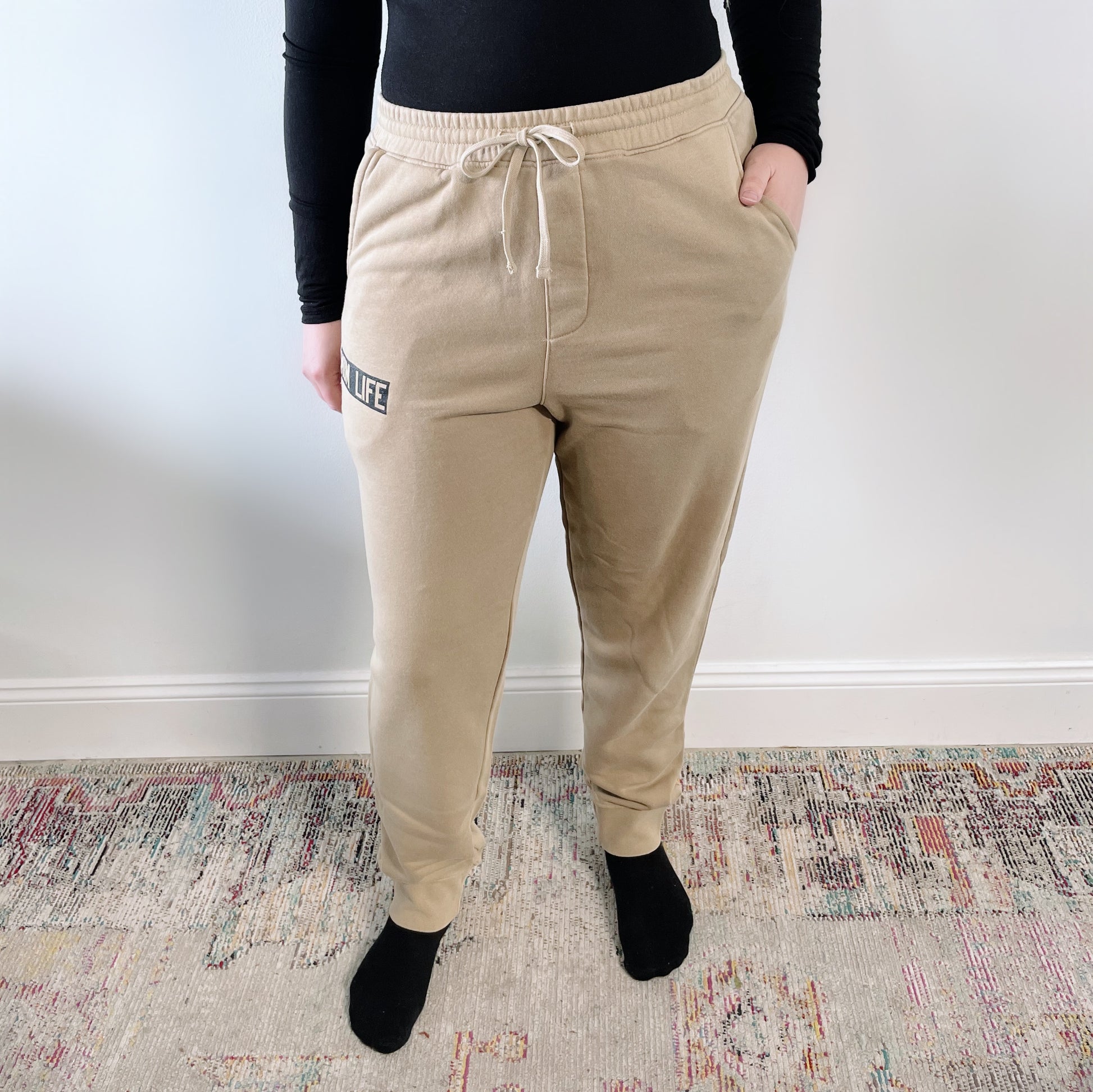 A person stands on a patterned rug against a white wall, hands in their pockets, wearing the Mom Life Joggers • Natural and a black top with black socks—perfect for cozy, laid-back comfort.
