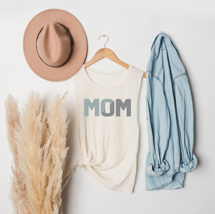 A beige hat, the MOM sliced • Dusty Scoop Tank—a sleeveless cream shirt with a relaxed fit—and a light denim shirt hang on hooks. Pampas grass stands beside them against a white wall.