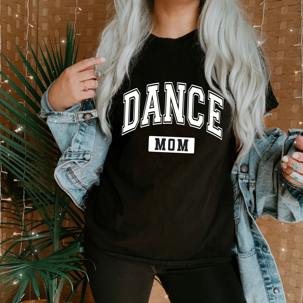 A person with long, wavy, silver hair wears a unisex Dance Mom • Tee from the Sports Mom Collection and a denim jacket, standing in front of a plant and brown woven background with string lights.