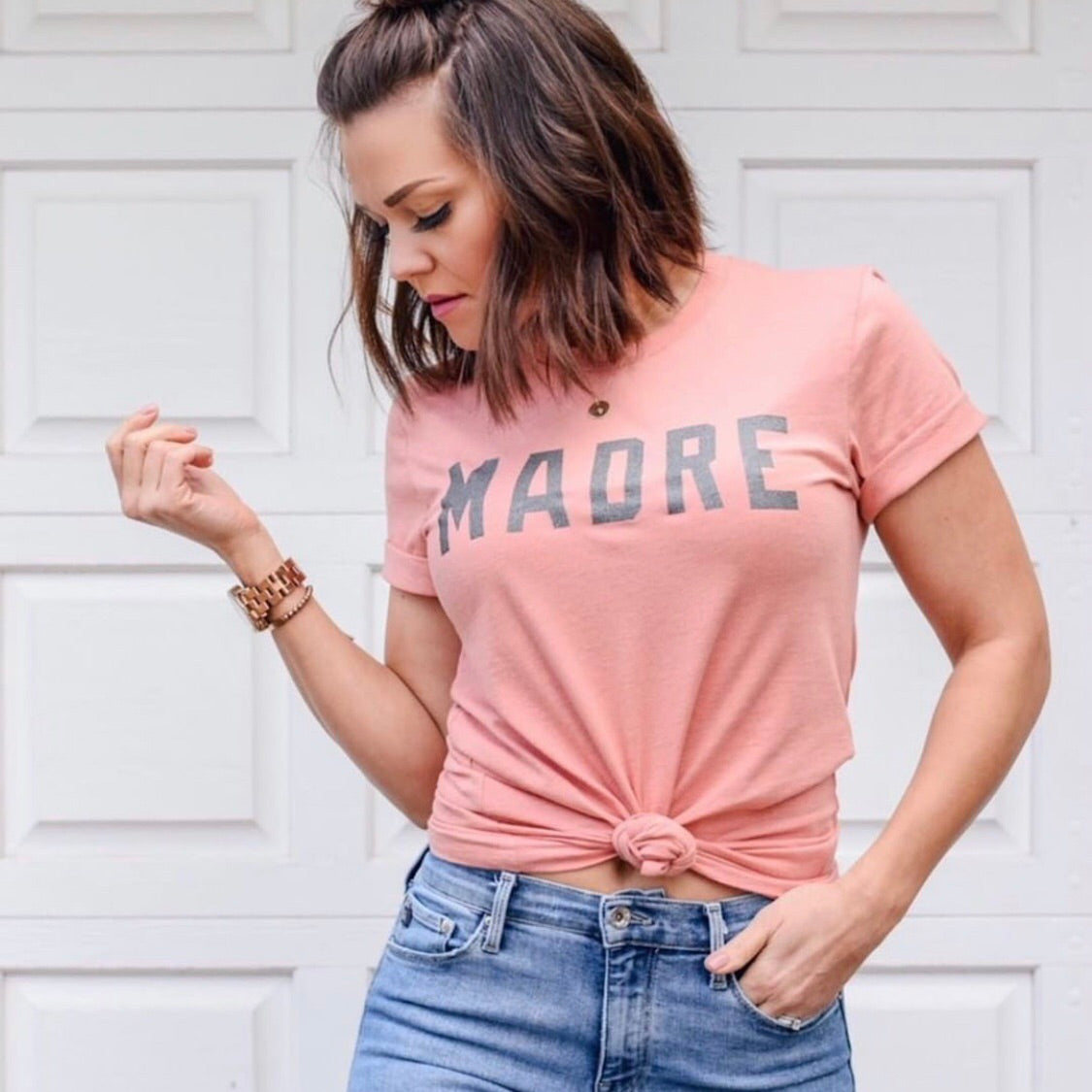 A woman in a Spring Coral Madre Tee stands before a white garage door, looking down and to the side, one hand raised and the other in her jeans pocket.