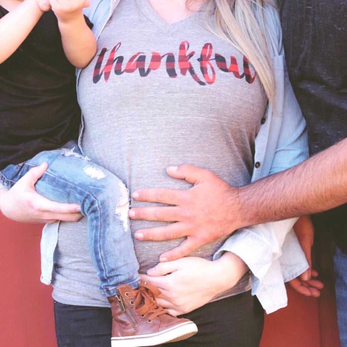 A pregnant woman in a Thankful Plaid • Tee holds a child, while an adult’s hand rests gently on her belly, capturing a warm family moment.
