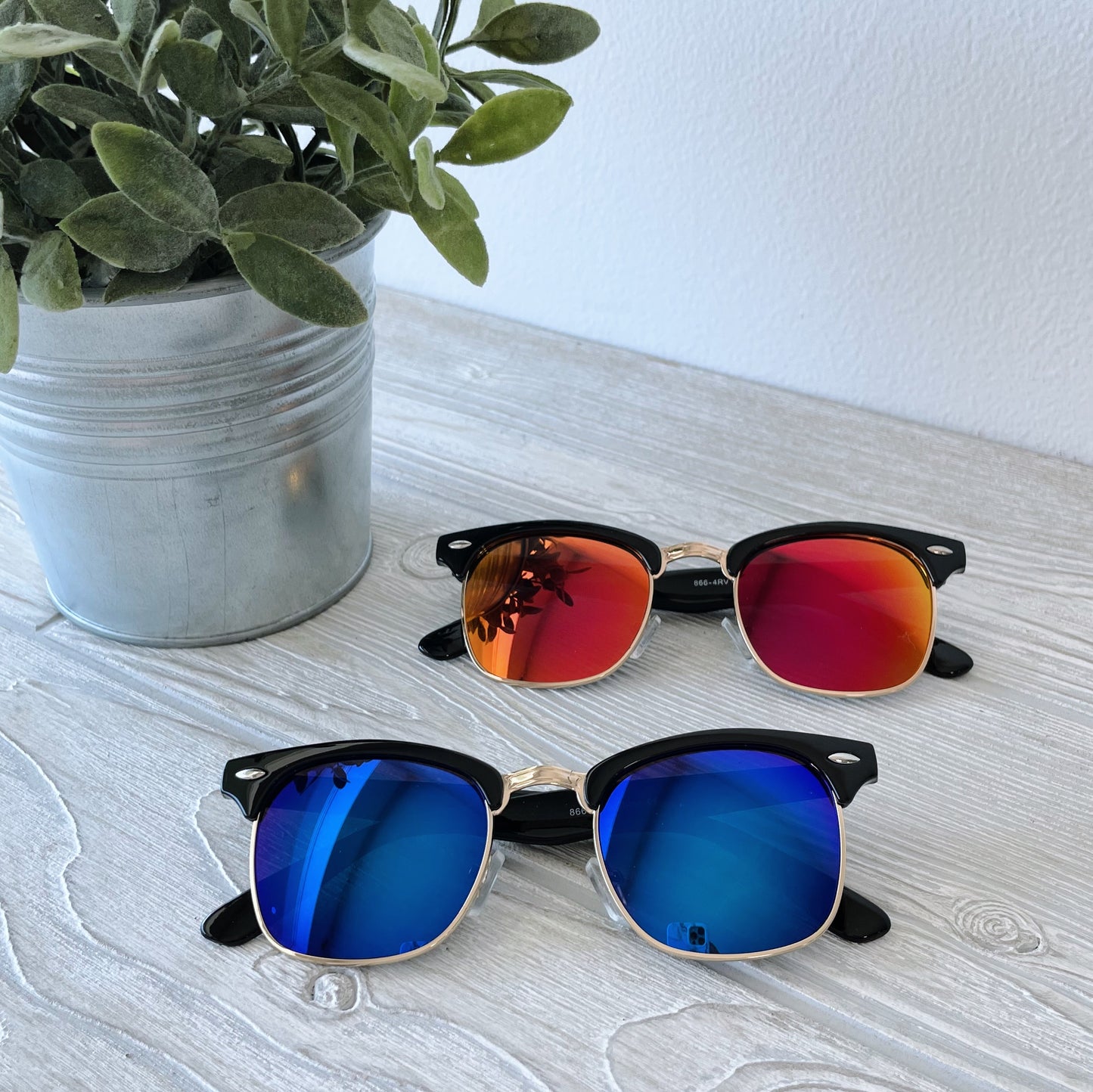 Two pairs of Parker • Adult Sunglasses with polarized mirrored lenses—one red-orange, one blue—sit on a light wooden surface next to a potted plant. Blue-light filtering makes these stylish shades both trendy and functional.
