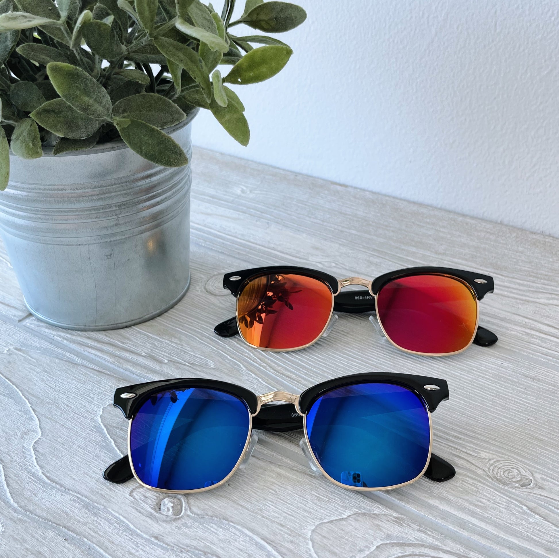 Two pairs of Parker • Adult Sunglasses with polarized mirrored lenses—one red-orange, one blue—sit on a light wooden surface next to a potted plant. Blue-light filtering makes these stylish shades both trendy and functional.