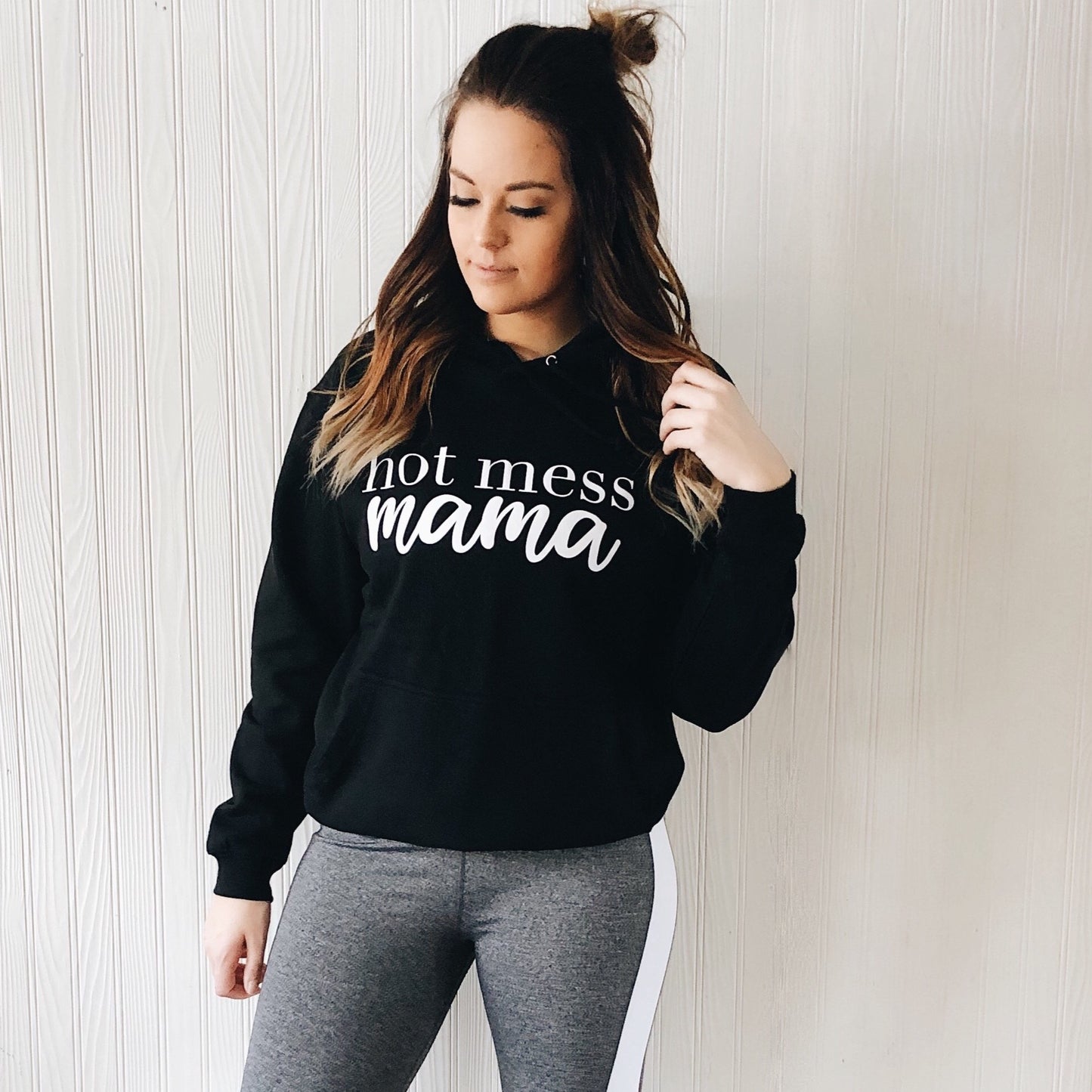 A woman with long hair stands against a white textured wall, wearing the Hot Mess Mama Black Hoodie and gray leggings with white side stripes for a cool monochrome look.