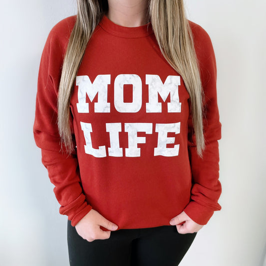 A woman poses against a light background wearing the MOM Marble • Brick Pullover, a red unisex sweatshirt featuring bold white MOM LIFE text on the front.