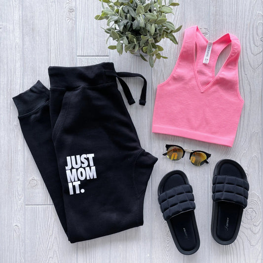Flat lay of casual loungewear: Just Mom It • Black Joggers, pink sleeveless crop top, black slide sandals, sunglasses, and a small green potted plant displayed on a light wood floor.