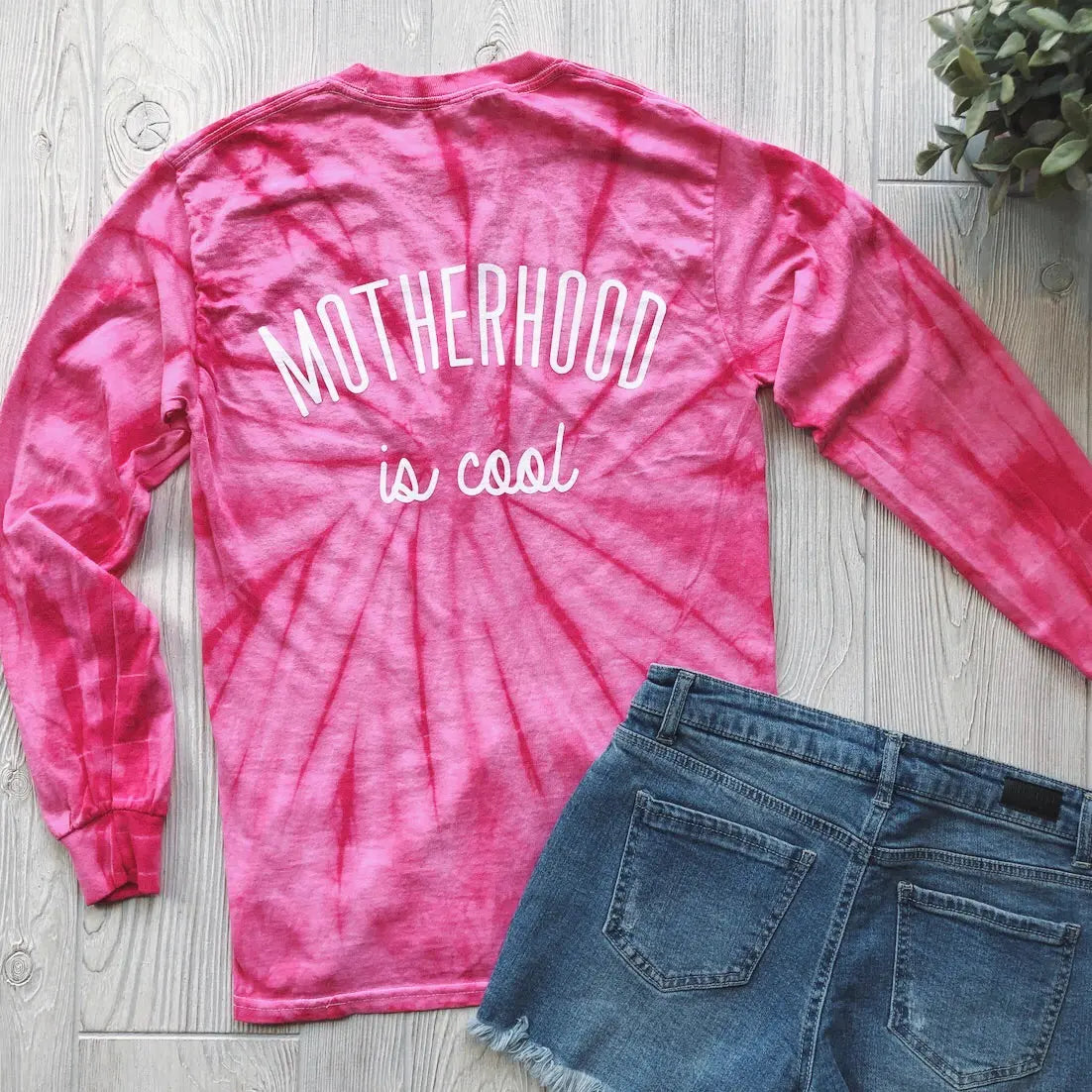The “Motherhood is Cool • Pink Longsleeve Tie-Dye” tee features a Motherhood design on the back and is shown flat on a light wooden floor with blue denim shorts and a potted plant.