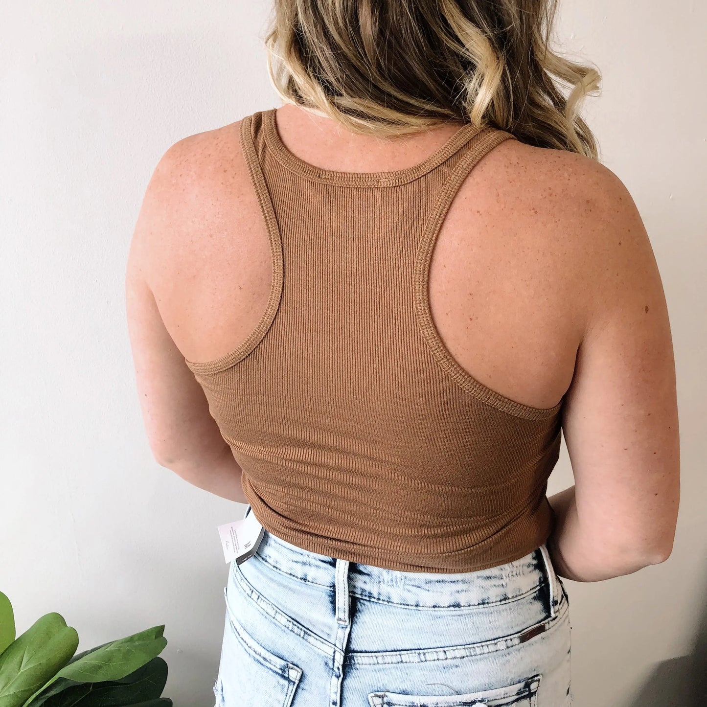 A woman with wavy blonde hair is seen from the back, wearing the New Basic Racerback Tank in brown with light-washed high-waisted jeans. She stands against a plain light wall, with part of a green plant visible. More colors available!.