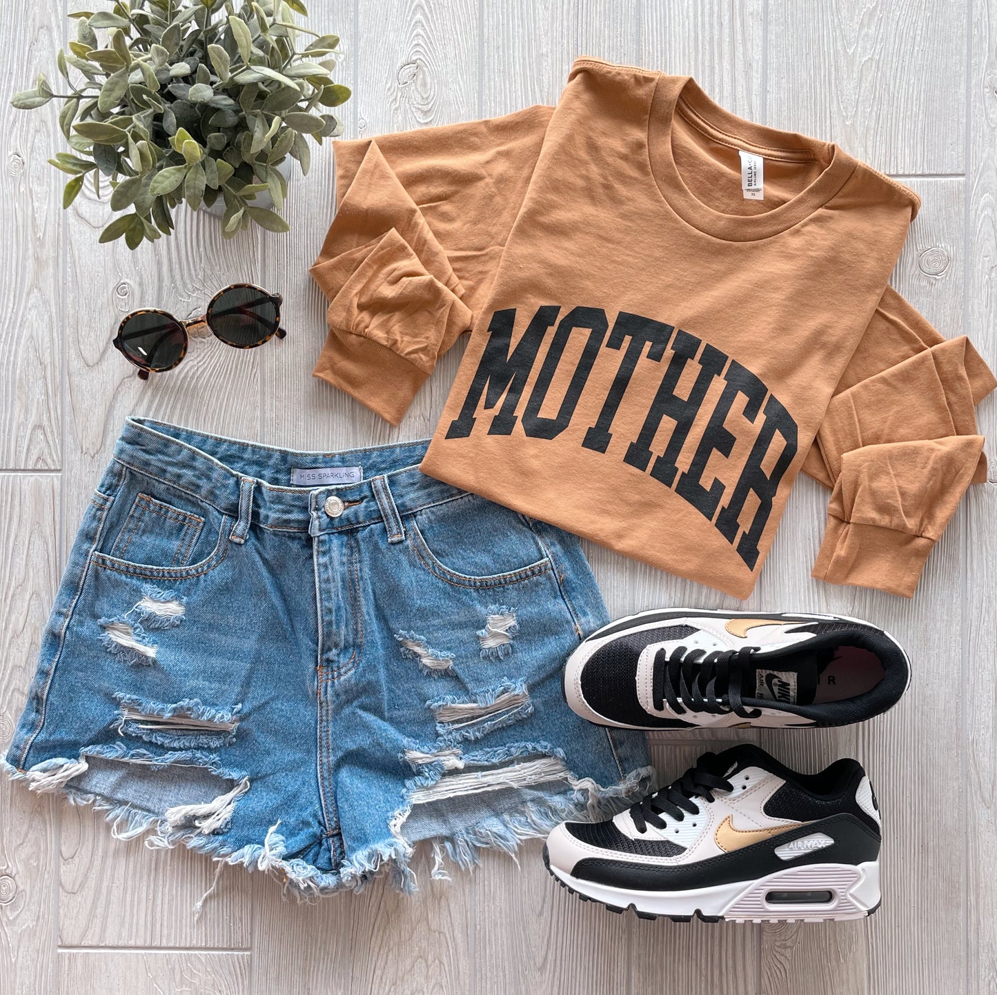 A flat lay of casual clothes featuring the MOTHER • Caramel Long Sleeve, distressed denim shorts, black and white Nike sneakers, round sunglasses, and a small potted plant on a light wood floor.