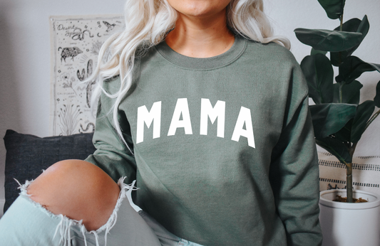 A person with long, light hair wears the MAMA • Military Green Pullover indoors, surrounded by a plant and wall art, with one knee in ripped jeans showing.