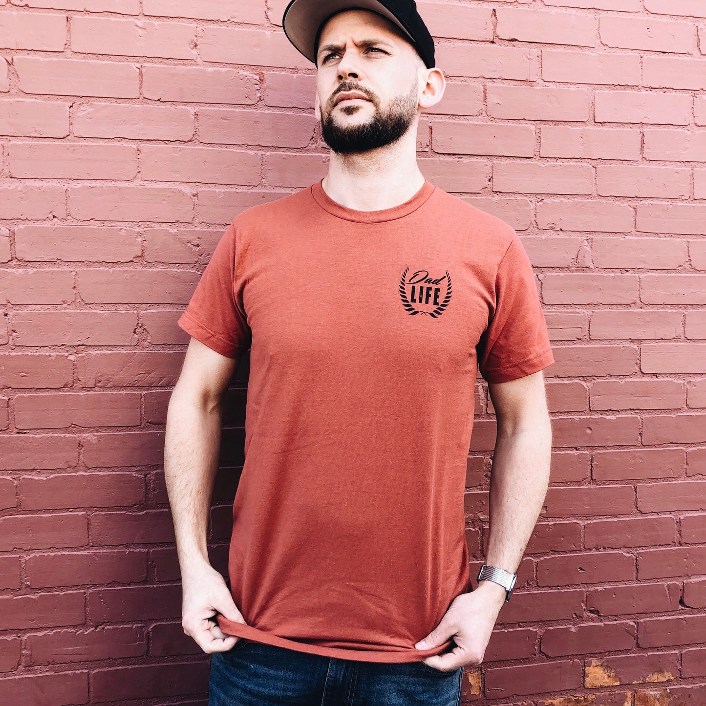 A bearded man in a Dad Life Left Chest • Rust Tee and black cap stands by a pink brick wall, gazing ahead and holding the hem of his pre-shrunk ring-spun cotton shirt featuring Life with a laurel graphic on the chest.