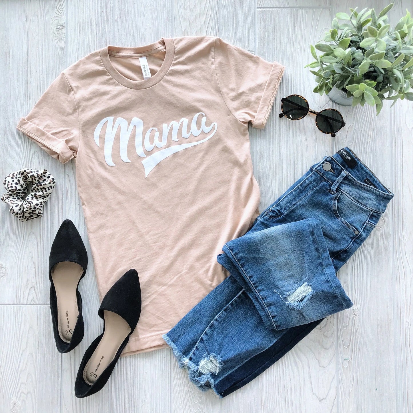 Flat lay of the Mama Sporty • Tan Tee paired with blue distressed jeans, black pointed flats, a scrunchie, round sunglasses, and a small potted plant on a light wood floor. The perfect comfy mom tee!.