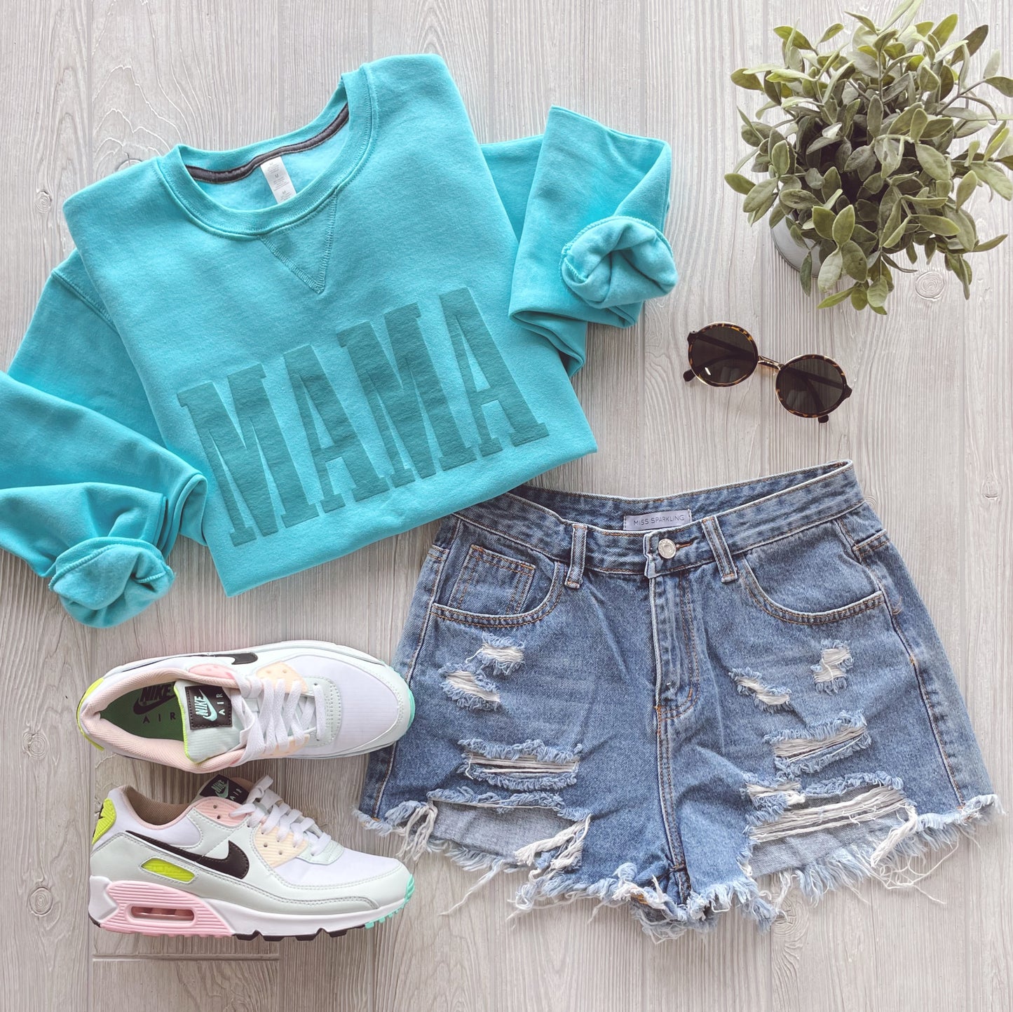 Flat lay of the MAMA • Aqua Tonal Pullover, distressed denim shorts, white Nike sneakers with colorful details, round sunglasses, and a potted plant on light wood. This comfy unisex sweatshirt delivers effortless style.
