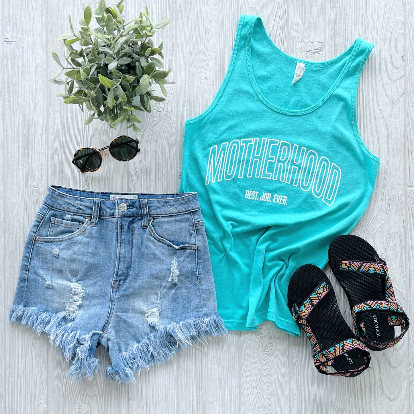 Flat lay of the Best. Job. Ever. • Unisex Tank Top in turquoise with “MOTHERHOOD Best. Job. Ever.” text, paired with distressed denim shorts, black sunglasses, patterned sandals, and a small green potted plant on a light wood background.