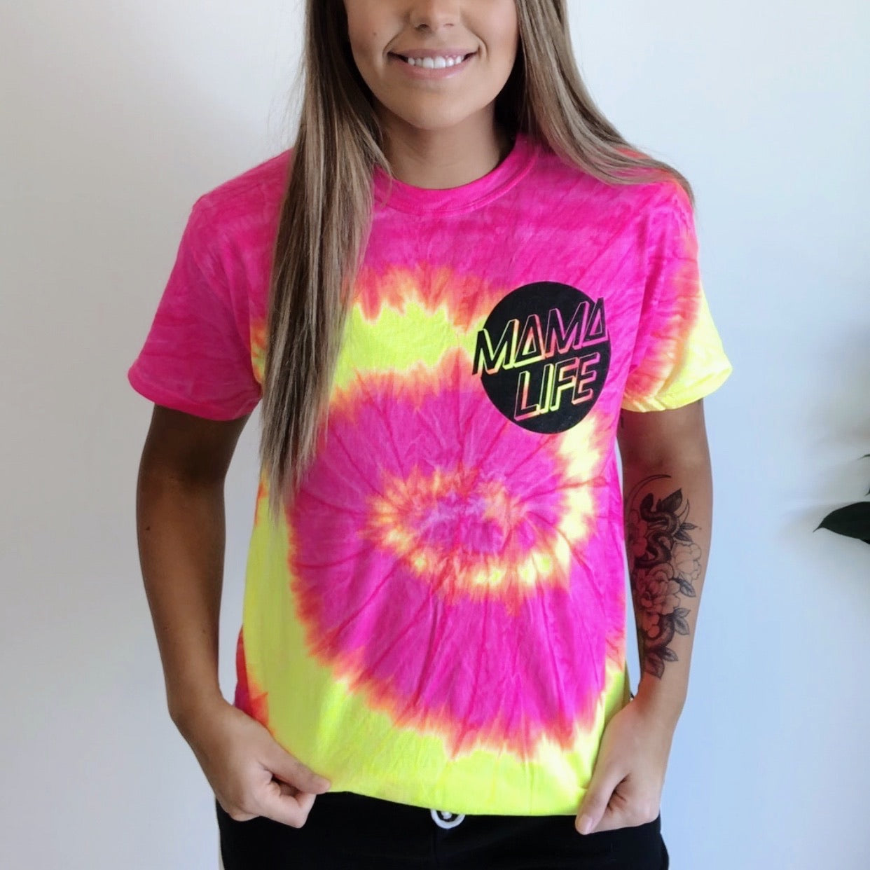 A smiling woman wears the Fluorescent Skater Mama • Tee Shirt—a vibrant Santa Cruz pink and yellow tie-dye top with MAMA LIFE in a bold black circle on the chest—showing off her left forearm tattoo.