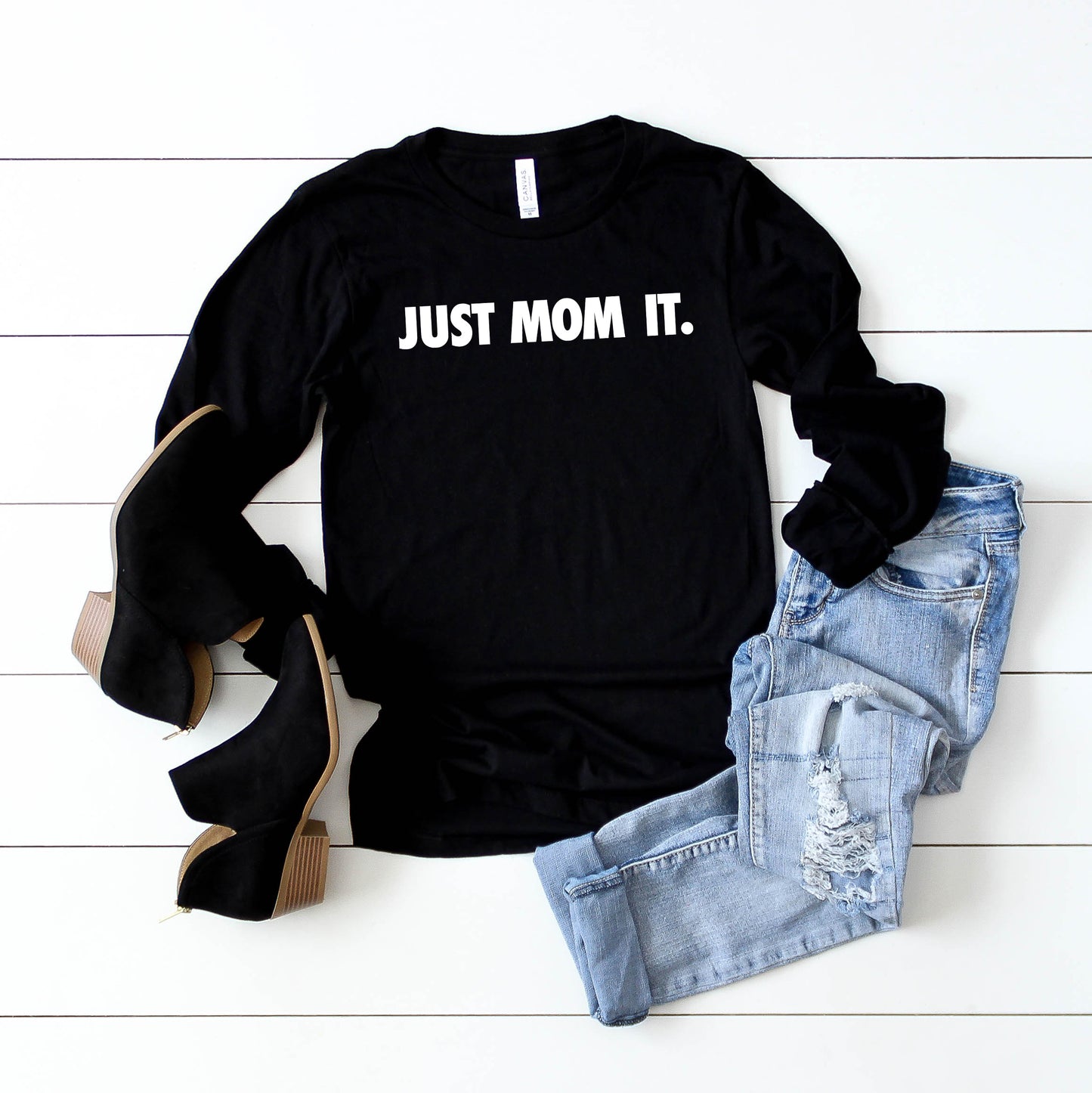 Flat lay of the Just Mom It • Black Long Sleeved tee in black with white lettering, styled with ripped light blue jeans and black heeled ankle boots on a white wooden background. Unisex sizing ensures a comfy fit for everyone.