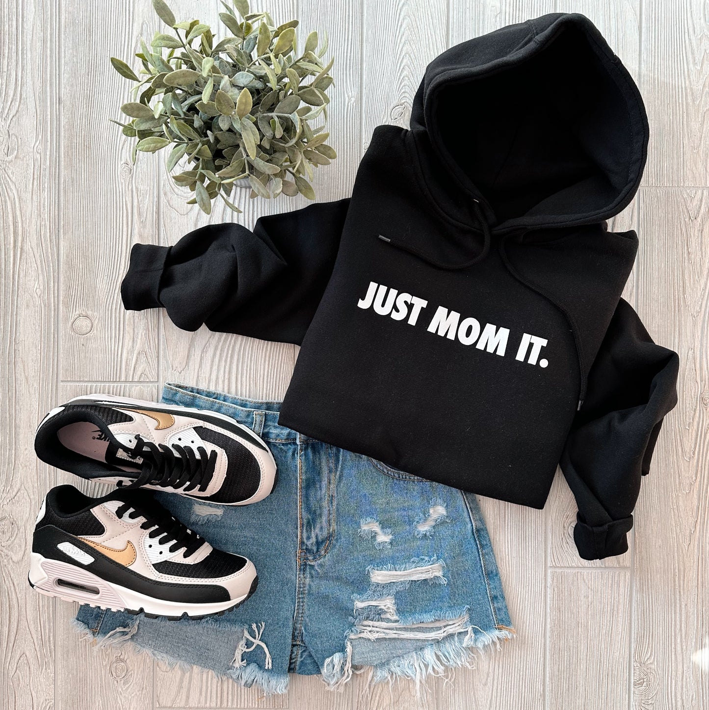 A flat lay of the Just Mom It • Black Hoodie, unisex fit, paired with distressed denim shorts, black and white sneakers with gold accents, and a green potted plant on a light wooden floor.