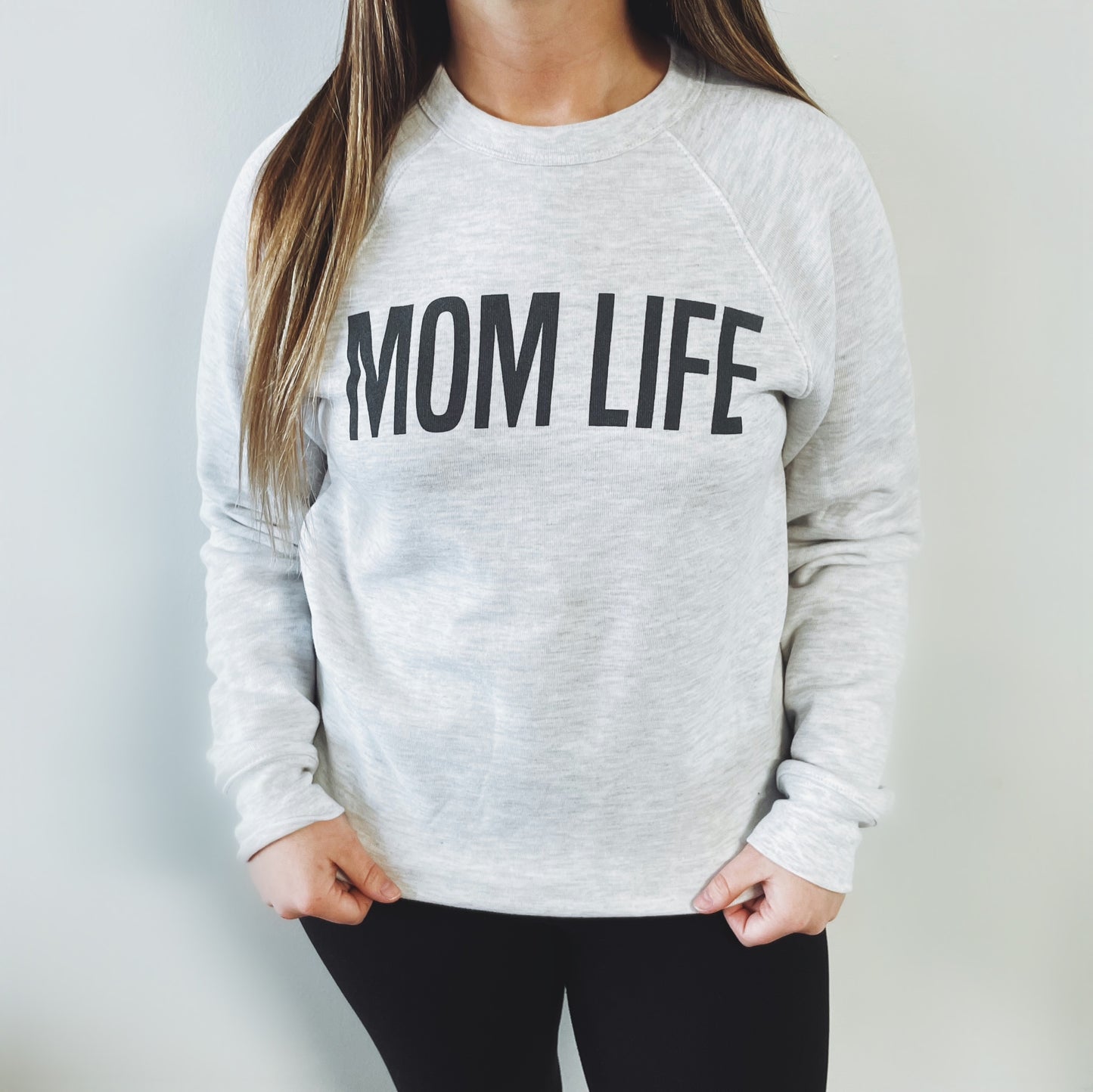 A woman stands against a light wall wearing the MOM LIFE • Ash Pullover, featuring bold black lettering and a unisex fit for comfortable style. She pairs it with black leggings; her face is not visible.