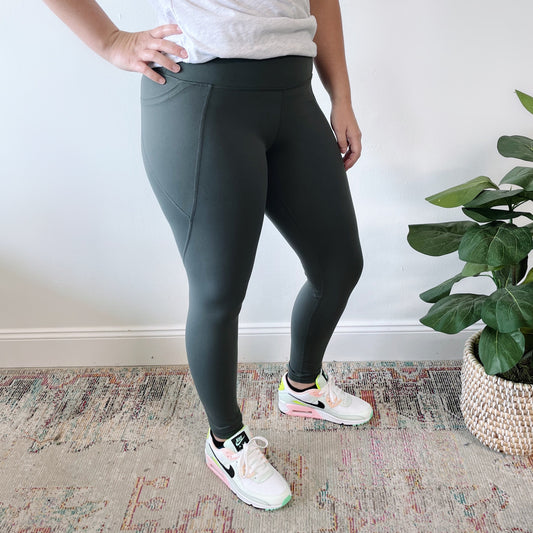 Someone wearing Ivy Angle Pocket Legging in dark green, featuring tummy control and side pockets, pairs them with a light gray top and pastel-accented white sneakers while standing on a patterned rug by a potted plant against a white wall.