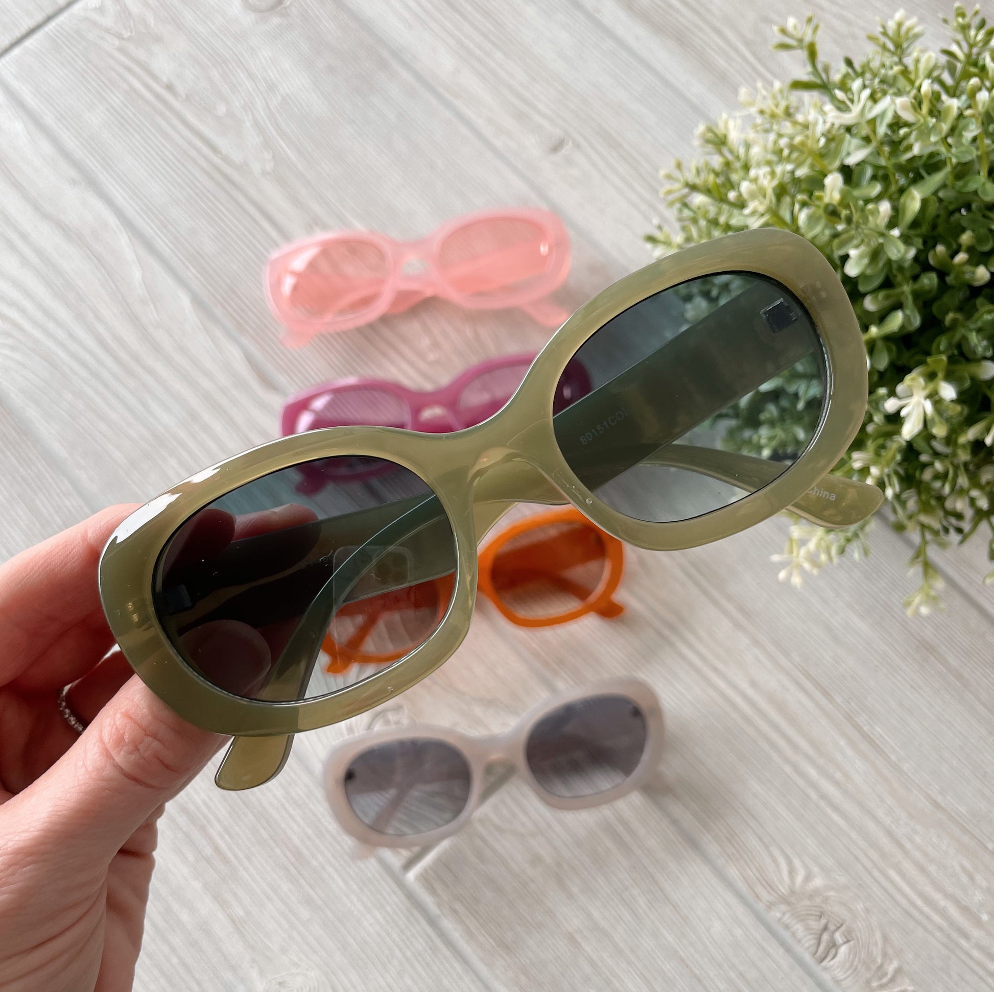 A hand displays olive-green Poppy Sunglasses • Adult • More Colors! On the light wood below are four more pairs—pink, purple, orange, and beige—with a small green plant nearby. All sunglasses are adult size and UV protected.