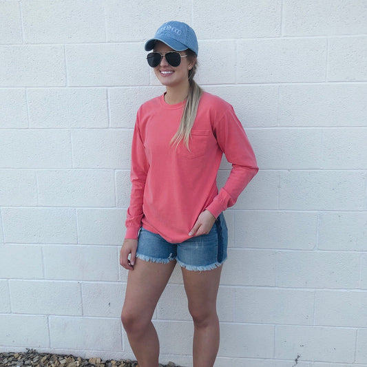 A woman stands against a white brick wall wearing the pink Motherhood • Pocket Long sleeve shirt, denim shorts, a blue baseball cap, and sunglasses. She smiles with one hand in her pocket.