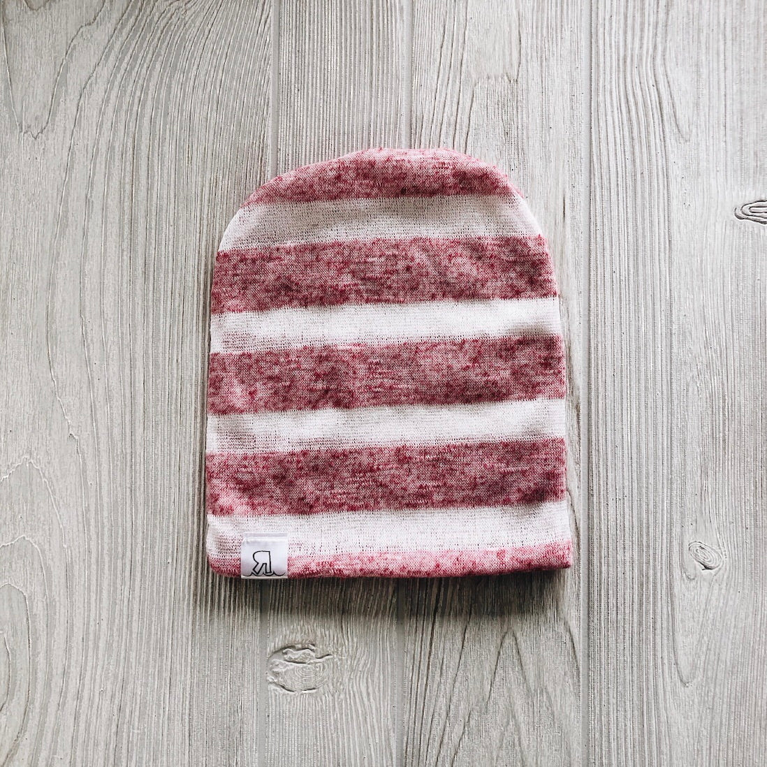 A Heather Pink Striped Beanie with horizontal pink and white stripes rests on a light gray wooden surface. A small white tag with a black logo accents the edge, showcasing its soft interlock knit construction.