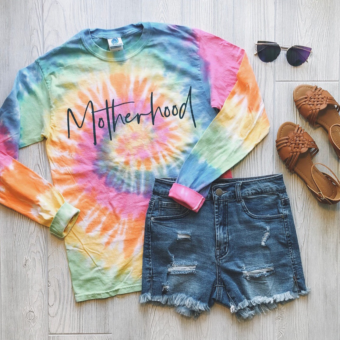 The Motherhood • Longsleeve Tie-Dye shirt with bold dark teal script is styled with distressed denim shorts, brown woven sandals, and black sunglasses, all arranged on a light wood floor.
