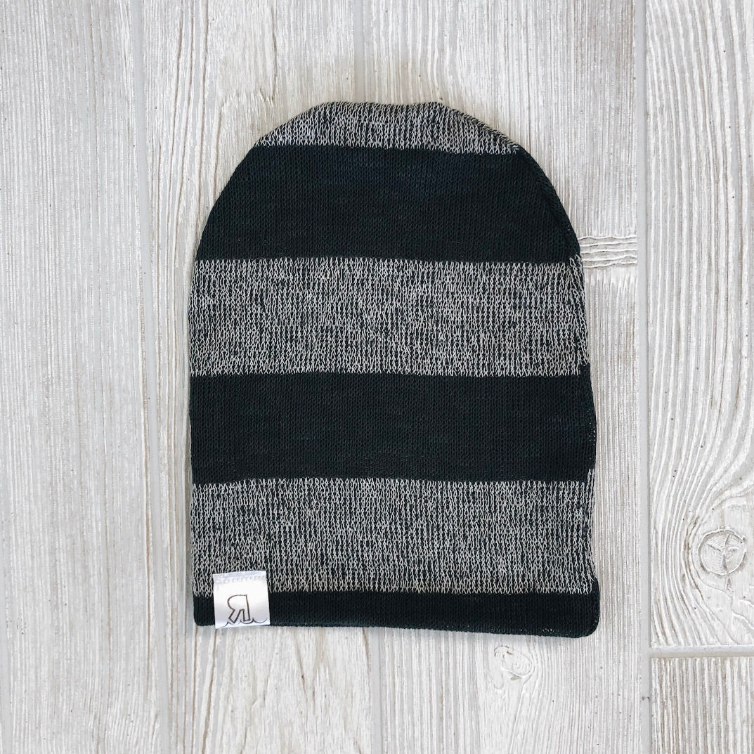 A Charcoal Stripe Beanie with black and gray stripes and a small white tag rests on a light wooden surface, highlighting its cute handmade style.