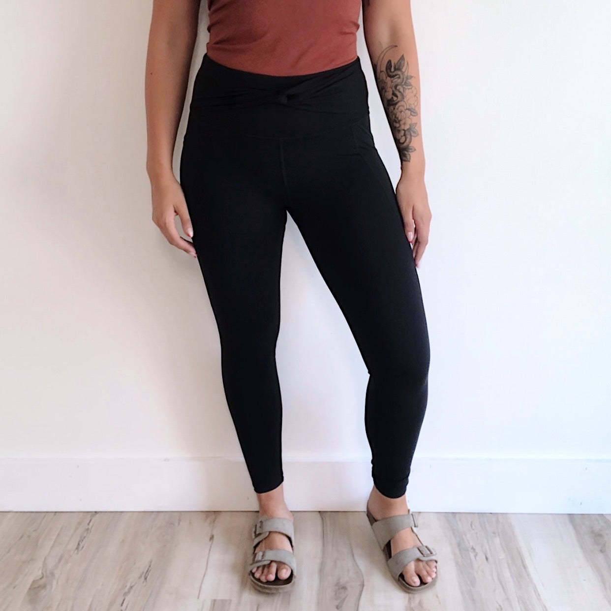 Standing indoors by a white wall, person wears a rust top, Twist Accent Highwaist Legging • 2869 with tummy control, and gray sandals. A floral tattoo decorates their left arm; the floor is light wood.
