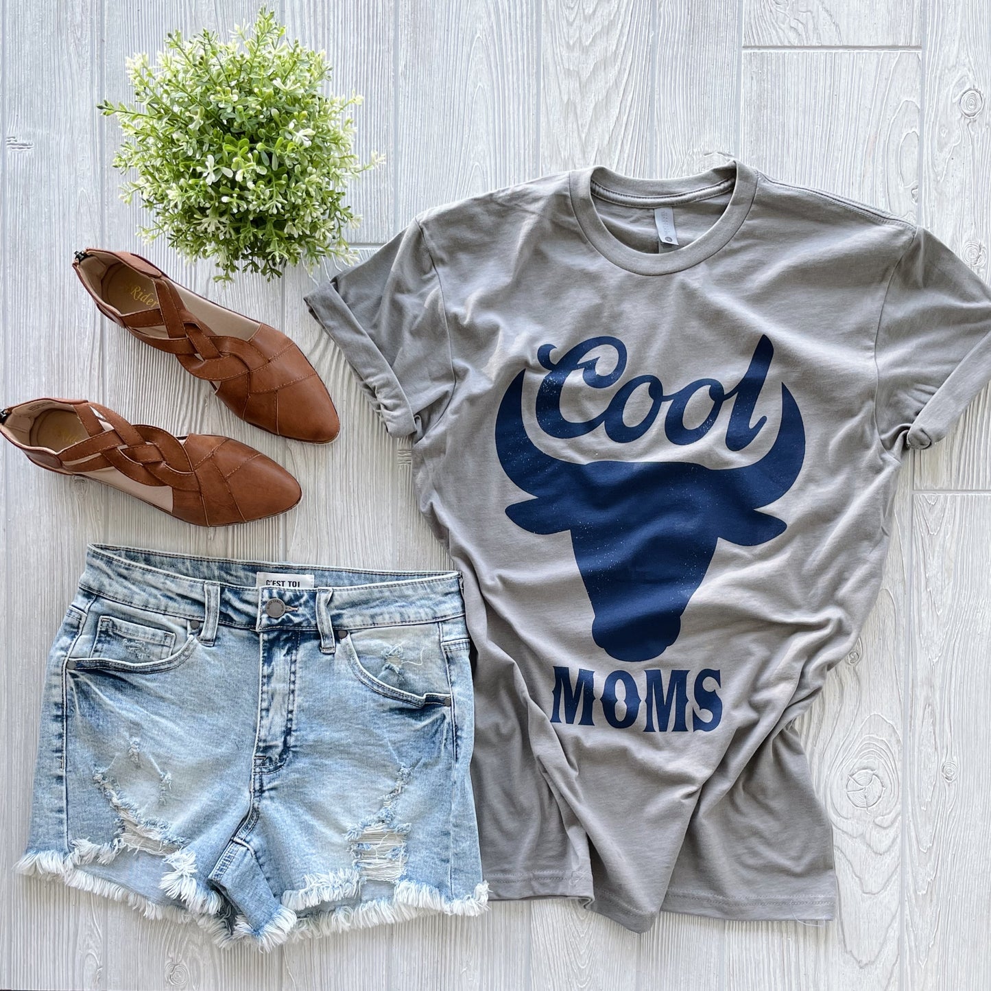 Flat lay featuring the Cool Moms • Stone Tee with bull graphic, paired with distressed denim shorts, brown woven flats, and a small green potted plant on a light wood floor. Unisex fit for effortless style.