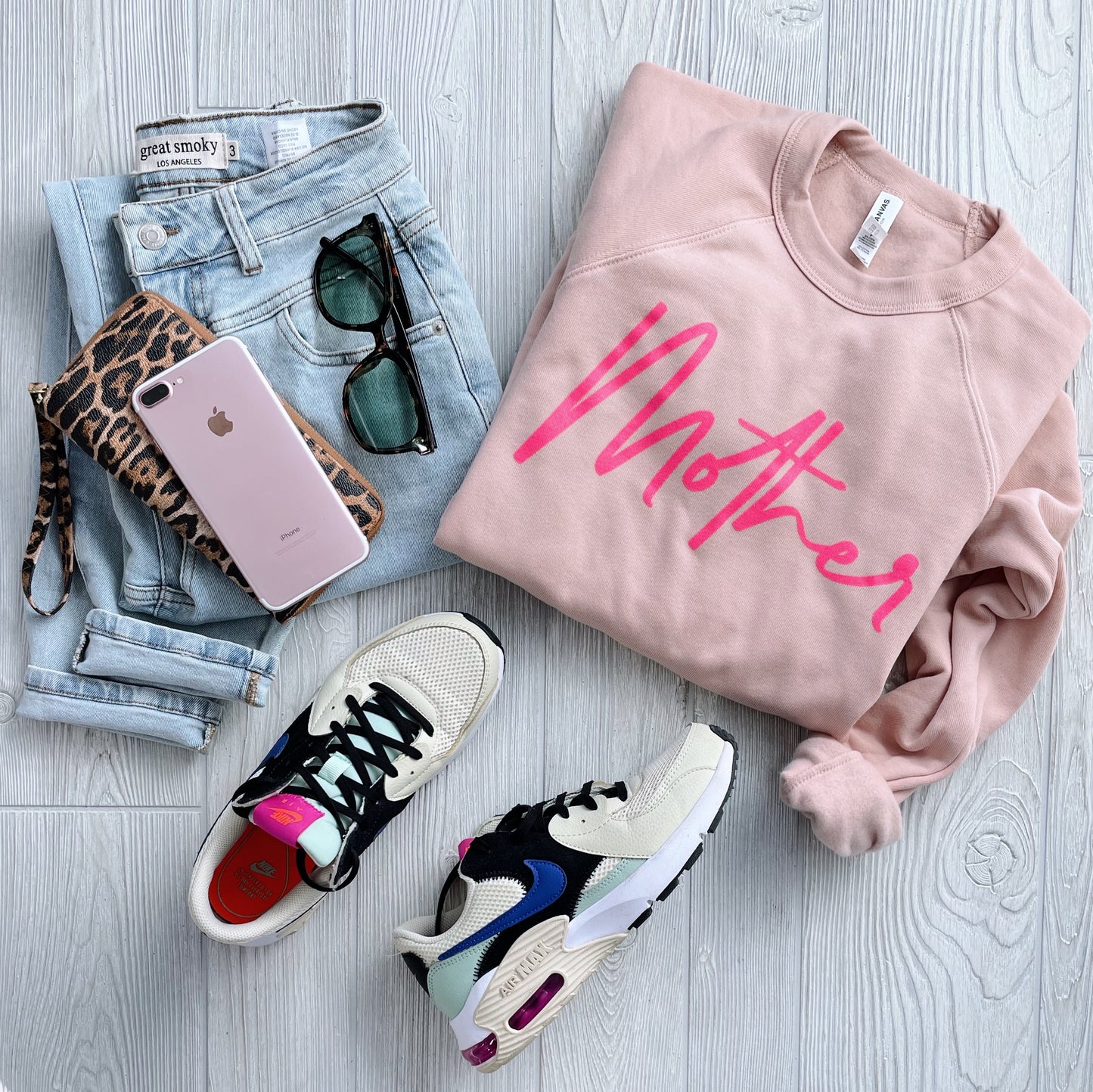 Flat lay of a casual outfit: light blue jeans, Mother • Peach Pullover sweatshirt, leopard print wallet, pink smartphone, sunglasses, and white, black, and pink sneakers on a light wooden floor.