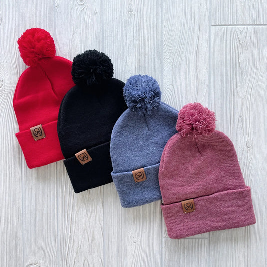 Four Pom Collection Beanies—red, black, blue, and mauve—are displayed on a light wood surface. Each hand-sewn beanie features a pom-pom and leather logo patch on the folded brim.