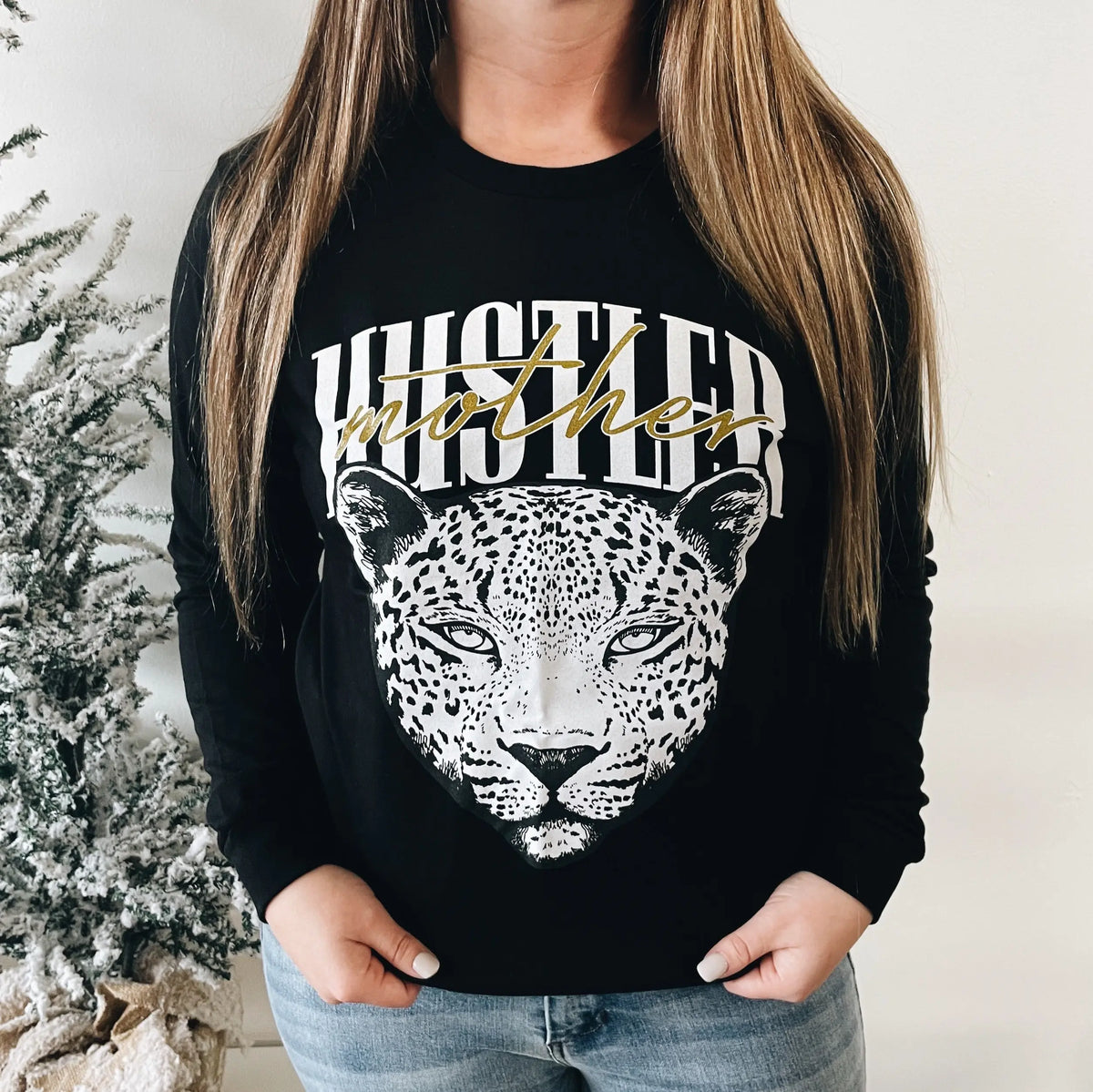 A woman in a black Cheetah Face • Long Sleeve tee with a bold leopard graphic and Mother Hustler text stands beside a frosted Christmas tree. This top is available in unisex sizing, making it ideal for a bold holiday statement.