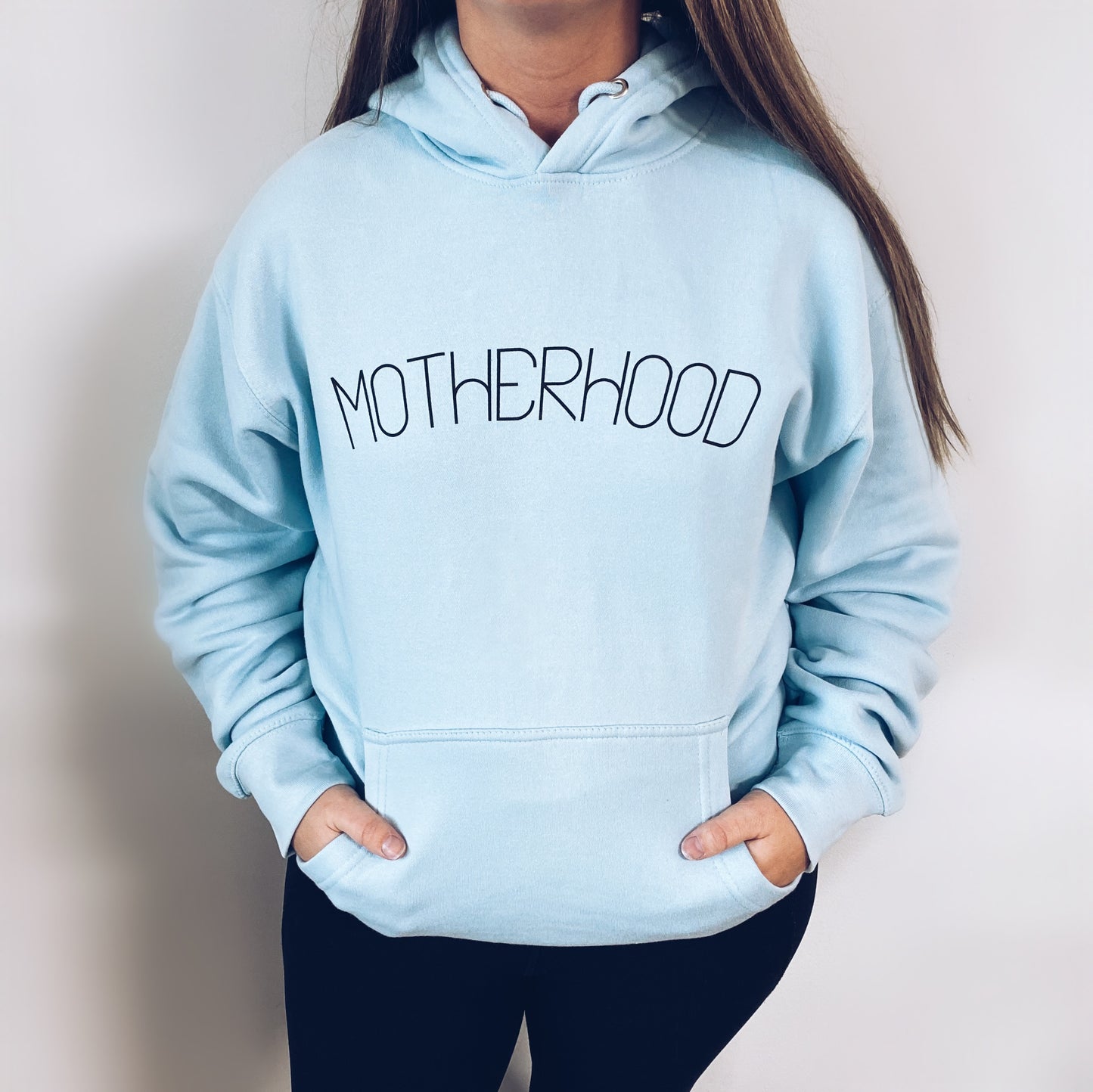 A woman stands against a white background, hands in the pocket of the Motherhood • Sky Blue Hoodie featuring a bold black Motherhood print on the front.