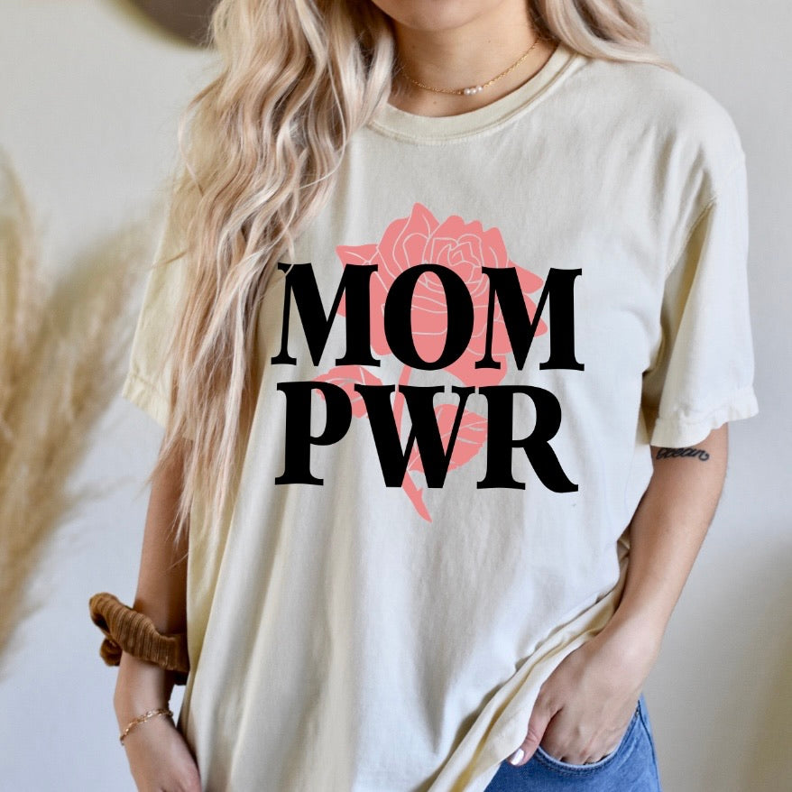 A woman with wavy blonde hair wears the MOM PWR • Natural Tee Shirt—a unisex beige top featuring bold black lettering and a pink rose design. She styles it with gold jewelry and keeps one hand in her pocket.