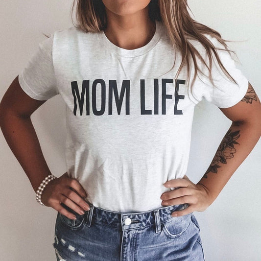 A woman stands with her hands on her hips wearing the MOM LIFE • Ash Tee, a light gray unisex shirt featuring bold black MOM LIFE text. She pairs it with ripped denim shorts and a beaded bracelet.