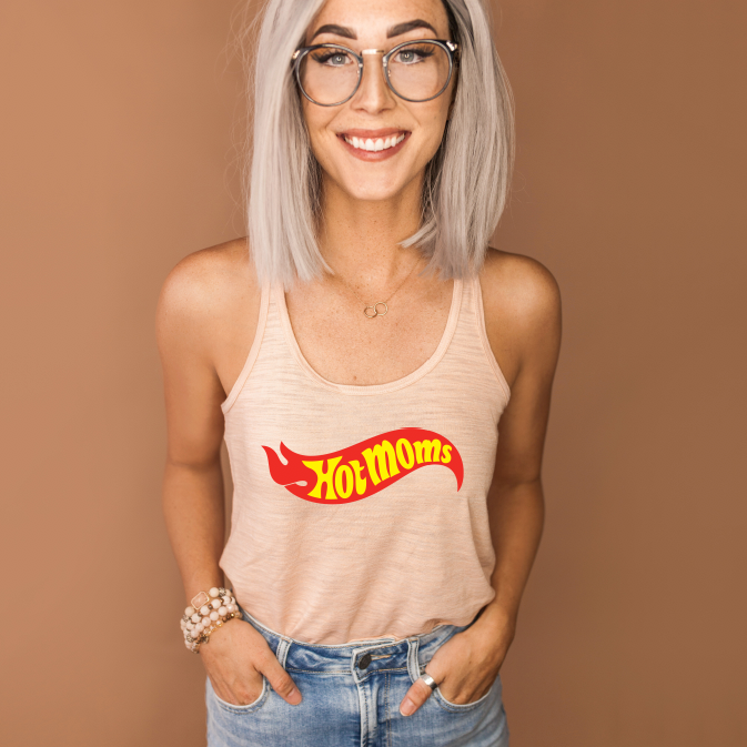 A smiling woman with light blonde hair and glasses wears the Hot Moms • Peach Tank, featuring bold red and yellow lettering. She has her hands in jean pockets and stands against a solid brown background.