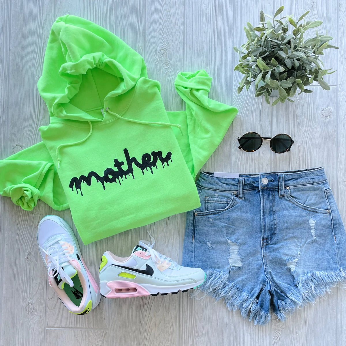 Flat lay of the Mother Slime • Lime Hoodie with unisex fit, distressed light denim shorts, white Nike sneakers with neon accents, round black sunglasses, and a small potted plant on a light wood background.