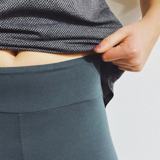 A person lifts their grey mesh top, revealing Wide-Waist Leggings in dark grey. The high-waisted style and soft brushed jersey show part of their midriff; only the torso and one hand appear against a plain white background. More colors available!.
