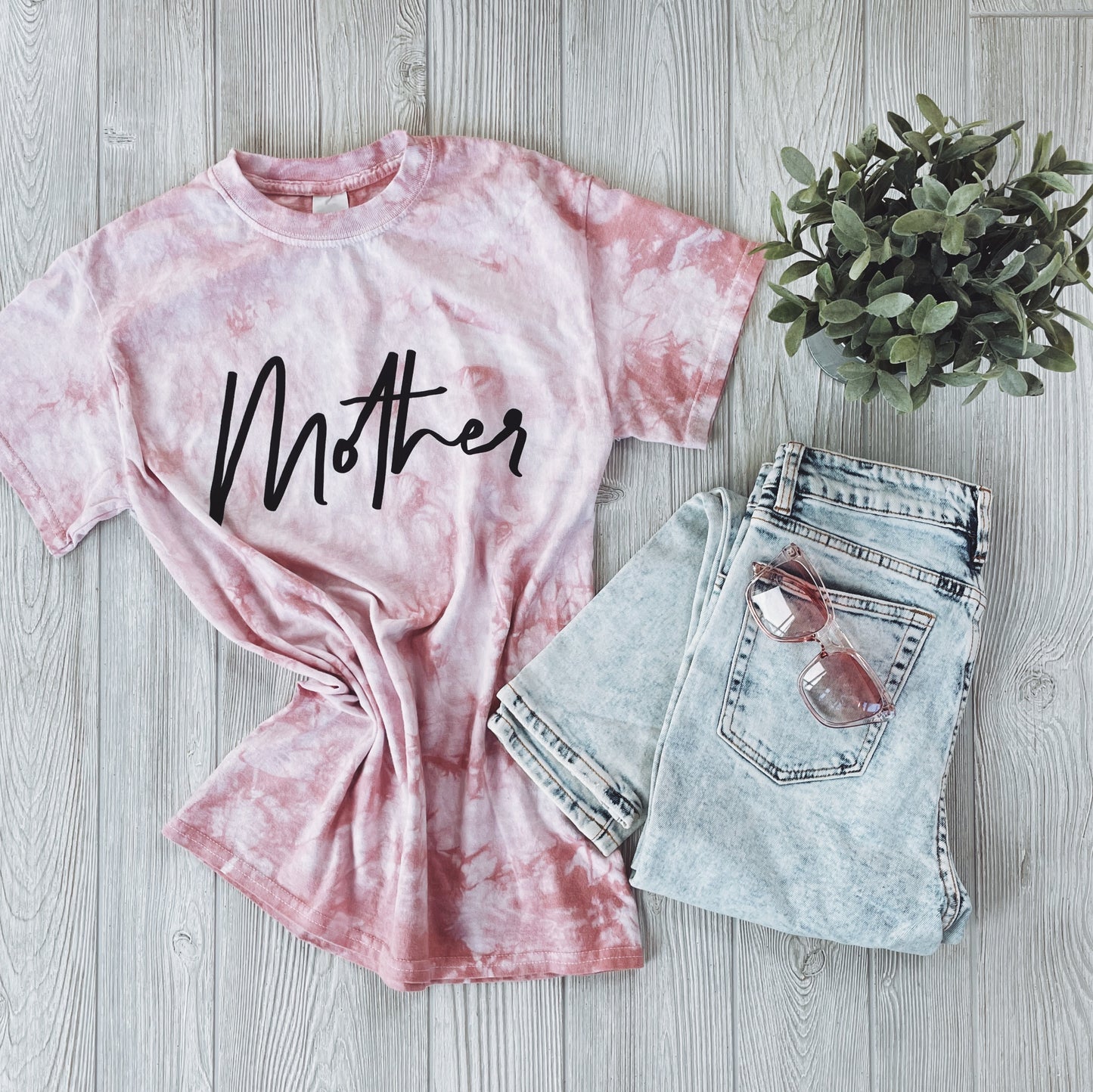 The Mother Tee • Rose Gold Tie Dye features black script on a relaxed, unisex fit. Styled with light-wash denim shorts, pink sunglasses, and a potted plant on a light wood floor—perfect for laid-back, casual outings.