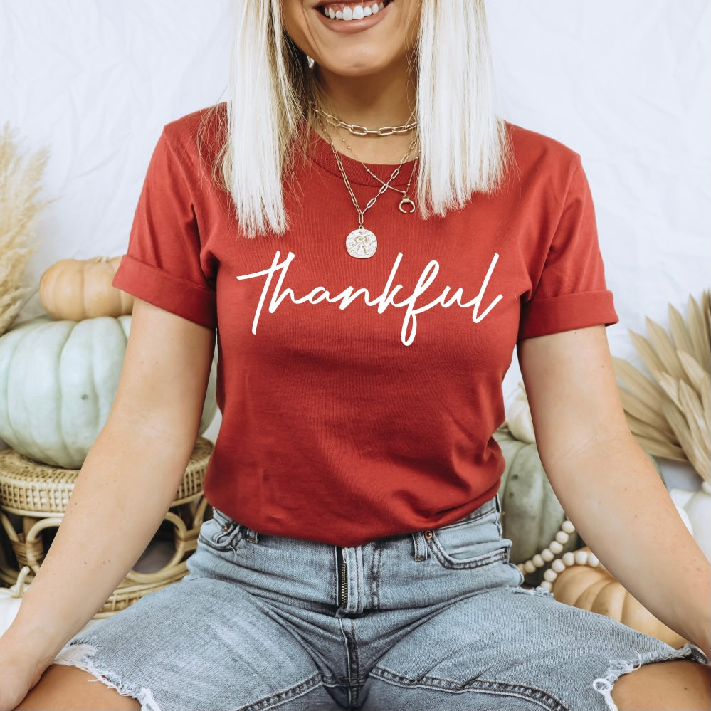 A smiling blonde woman wears the thankful • Rust Tee—a comfy red t-shirt with “thankful” in white script—paired with denim shorts, sitting cross-legged among neutral pumpkins and autumn decor.
