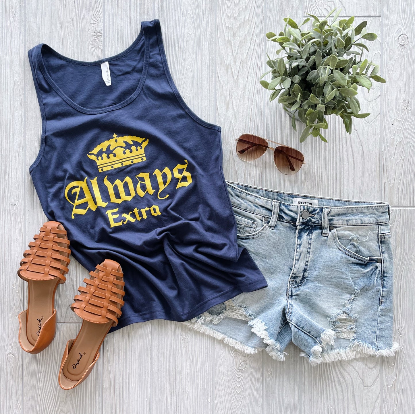 Flat lay of the Always Extra • Unisex Tank Top in navy with a yellow crown and “Always Extra” text, paired with distressed denim shorts, tan woven sandals, brown sunglasses, and a small plant on a light wood floor. Unisex fit.