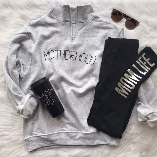 Flat lay of a gray unisex fit Motherhood • Quarter Zip Pullover, black MOM LIFE leggings, black travel mug, and brown sunglasses arranged on a white furry surface.