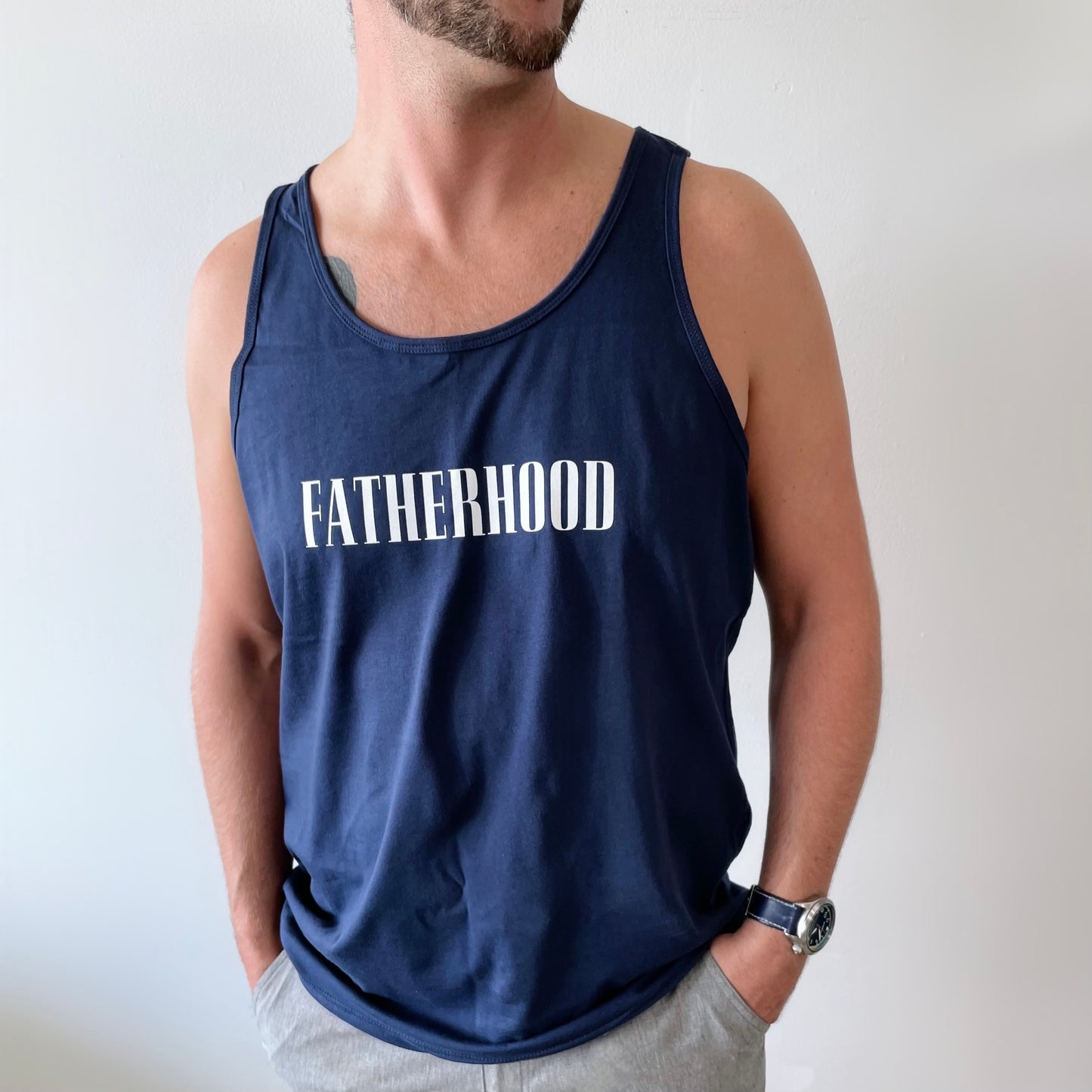 A man stands against a light background, hands in pockets, wearing our FATHERHOOD • Navy Tank from the unisex blanks collection with FATHERHOOD printed in bold white letters.