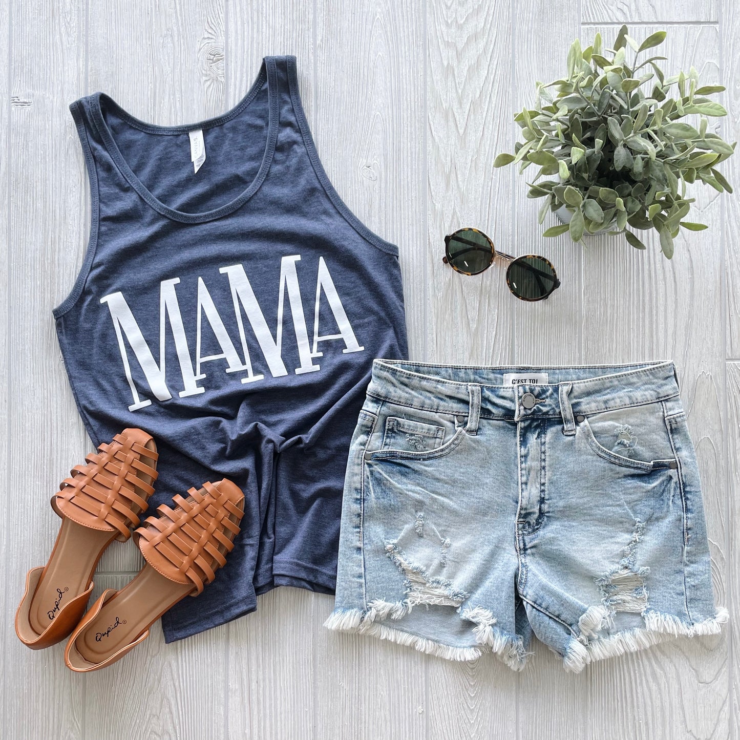 A flat lay displays the MaMa • Unisex Tank Top in navy with a white design, styled with distressed denim shorts, brown sandals, round sunglasses, and a small potted plant on a light wood background.