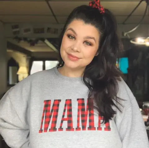 A woman with dark hair in a ponytail and a red plaid scrunchie smiles, wearing the MAMA • Plaid Print Pullover—this cozy gray unisex sweatshirt features bold red plaid MAMA letters, perfect for soft-lit days indoors.