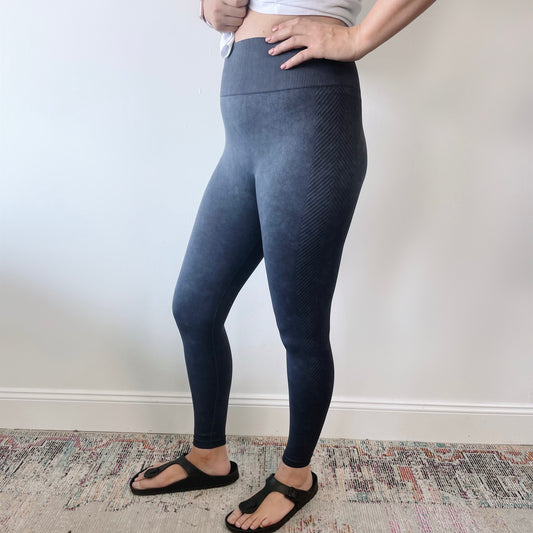 A person wearing Charley Mineral Wash Leggings • 8016—high-waisted, dark gray with tummy control—and black sandals stands indoors on a patterned rug against a plain white wall, hands resting on their waist.