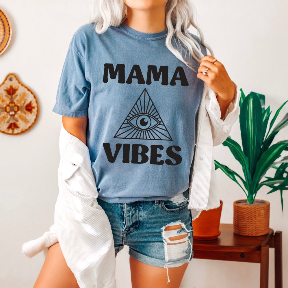 A woman with long, wavy hair wears the blue MAMA VIBES • Comfort Colors Tee featuring an all-seeing eye in a triangle, styled with a white open shirt and denim shorts. She stands near a leafy plant and a small wooden table.
