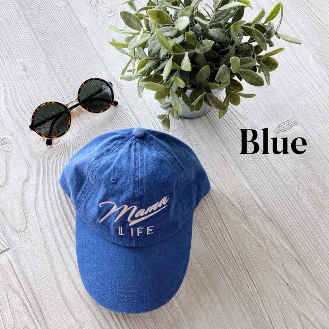 The Mama Life Hat in blue, featuring vintage white embroidery, is shown with round sunglasses and a potted plant on light wood. Blue appears in black on the right.